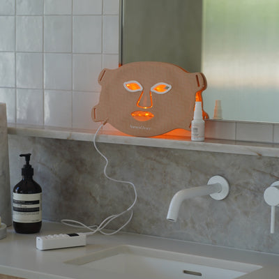 NursesChoice LED face mask lit on a bathroom vanity beside the Hyaluronic Acid Serum, stocked by Nurse Lisa at The Glow Co. Aesthetics, Gordon Park Brisbane