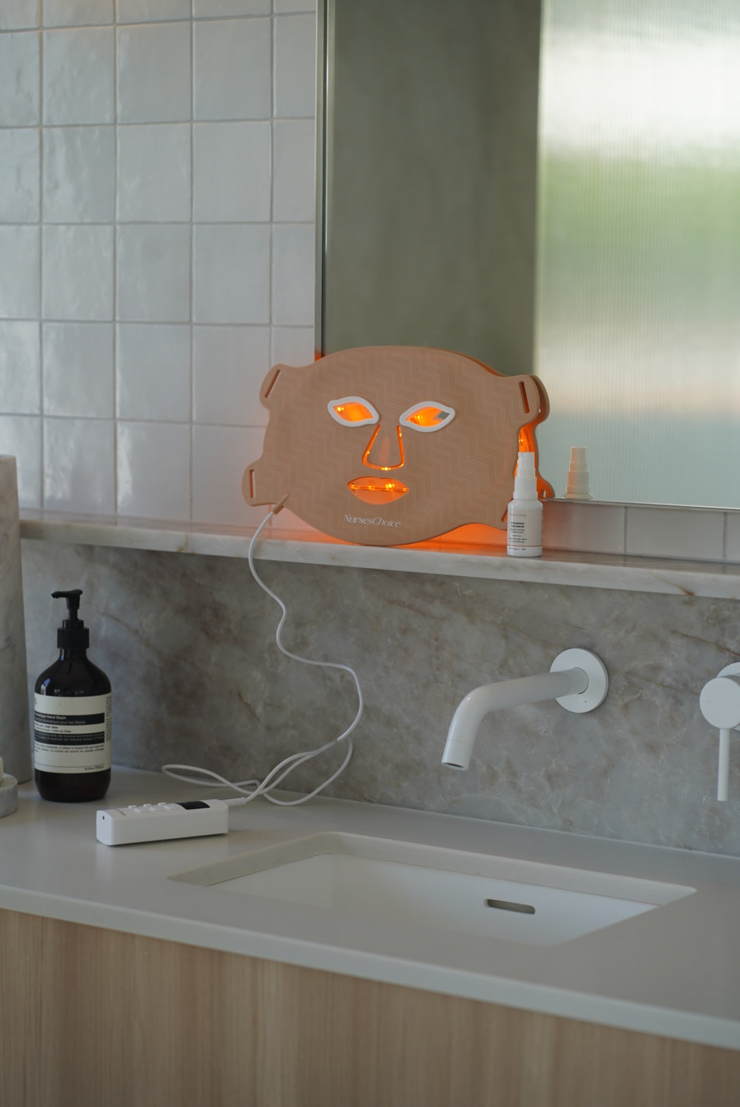 NursesChoice LED face mask lit on a bathroom vanity beside the Hyaluronic Acid Serum, stocked by Nurse Lisa at The Glow Co. Aesthetics, Gordon Park Brisbane